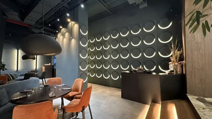 Dark gray geometric gypsum 3D wall panels with integrated LED lighting, creating a bold accent wall in a modern hospitality space. These premium plaster 3D panels from Kandes, a Swiss producer of high-quality 3D wall panels, enhance interior design with circular cut-out patterns, perfect geometry, and ambient lighting. Made from all-natural gypsum, A1 non-combustible, and eco-conscious, they provide seamless wall cladding for commercial and residential projects. Paintable for full customization.